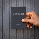 Wayfinder LET'S GO "FOREST BATH" B7 passport size notebook shown cover closed in front of a concrete wall. 